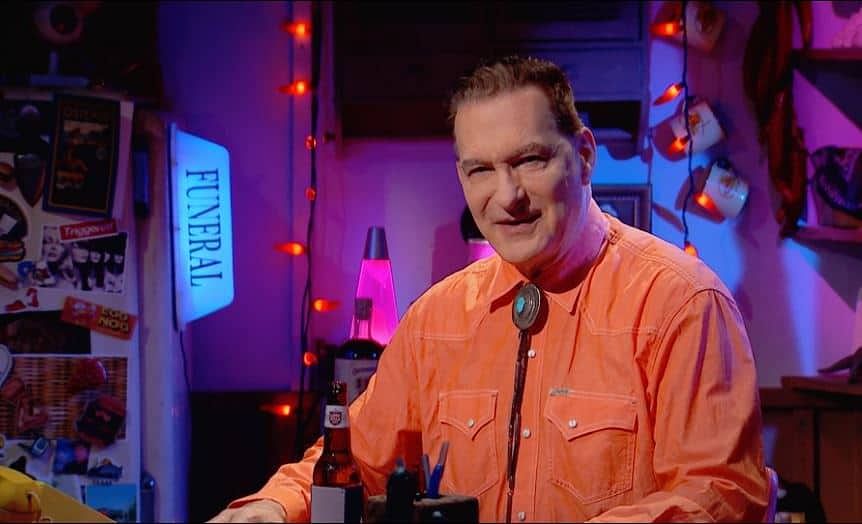 A man in an orange shirt sitting at a table while watching The Last Drive-In Season 1 Week 7 – A Girl Walks Home Alone at Night.