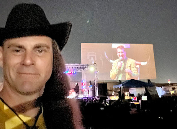 A man in a cowboy hat standing in front of a screen while wearing an auto draft shirt.