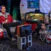 Joe Bob Briggs sits wearing a western shirt with Darcy the Mail Girl in black boots relax by a trailer with beer bottles, a cooler, TV, and fish tank. Behind them, an autograph sign reads "The Last Drive-In Never Dies" from The Last Drive-In with Joe Bob Briggs 2025