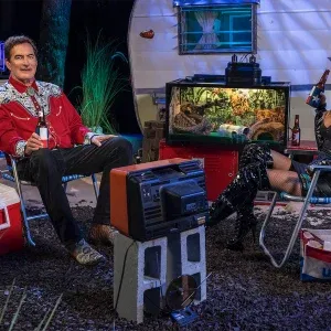 Joe Bob Briggs sits wearing a western shirt with Darcy the Mail Girl in black boots relax by a trailer with beer bottles, a cooler, TV, and fish tank. Behind them, an autograph sign reads "The Last Drive-In Never Dies" from The Last Drive-In with Joe Bob Briggs 2025